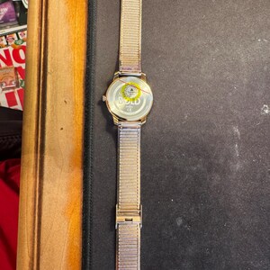 May include: A gold-toned wristwatch with a mesh band and a round face. The watch has a gold case and a silver clasp. The watch face has the word "BOLD" printed on it. The watch is lying on a black surface.