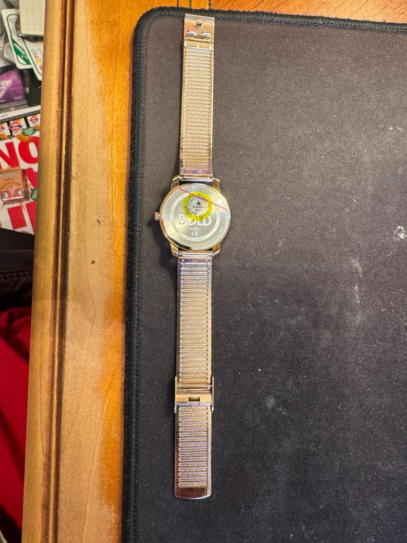 May include: A gold-toned wristwatch with a mesh band and a round face. The watch has a gold case and a silver clasp. The watch face has the word "BOLD" printed on it. The watch is lying on a black surface.