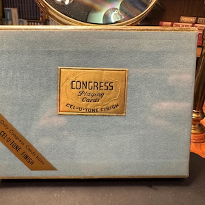 Könnte beinhalten: Eine Vintage-Spielkartenbox von Congress. Die Box ist hellblau mit goldenen Zierleisten und einem goldenen Etikett mit der Aufschrift "CONGRESS Playing Cards CEL-U-TONE FINISH". Ein goldenes Ecketikett lautet "Only Congress Cards have CEL-U-TONE FINISH."
