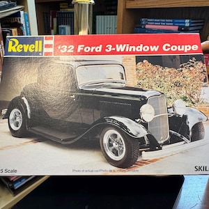 Puede incluir: Una caja de kit de modelo Revell para un '32 Ford 3-Window Coupe. La caja presenta un coche vintage negro con ruedas y faros cromados. El texto en la caja incluye 'Revell', '32 Ford 3-Window Coupe', '1:25 Scale' y 'SKILL 2'.