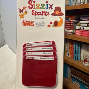 Op de afbeelding: Een Sizzix Sizzlits stansvormenset voor herfstthema knutselen. De verpakking bevat stansvormen voor een hoorn des overvloeds, de zin "Give Thanks", een pelgrimshoed en bladeren en eikels. De verpakking is wit met rode accenten.