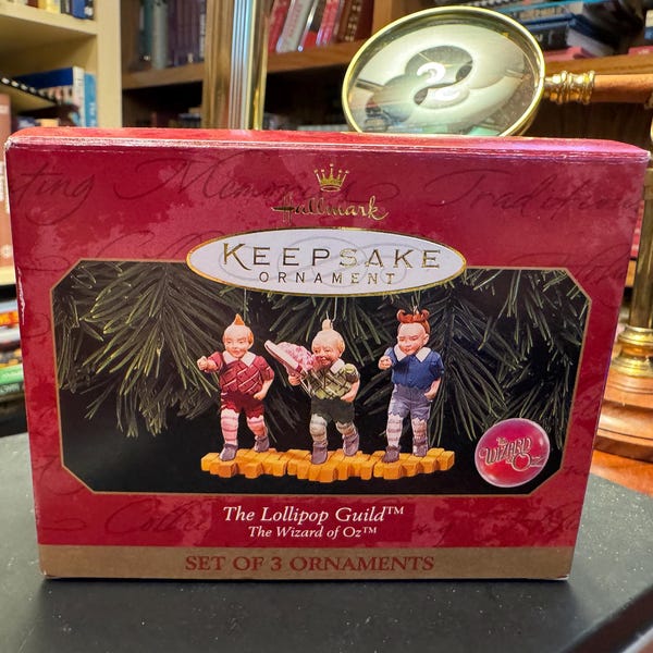 May include: A Hallmark Keepsake Ornament box featuring three Lollipop Guild figures from The Wizard of Oz. The box is red with gold accents and the text "The Lollipop Guild" is visible. The set includes three small figurines.