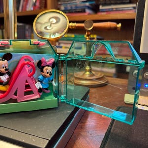 May include: A clear, turquoise plastic toy house with Mickey and Minnie Mouse figurines and a pink slide inside. The house is open, revealing the figures and play structure. The background includes a desk and a magnifying glass.