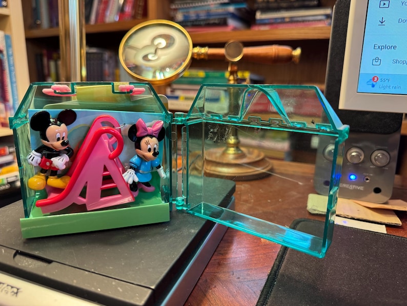 May include: A clear, turquoise plastic toy house with Mickey and Minnie Mouse figurines and a pink slide inside. The house is open, revealing the figures and play structure. The background includes a desk and a magnifying glass.