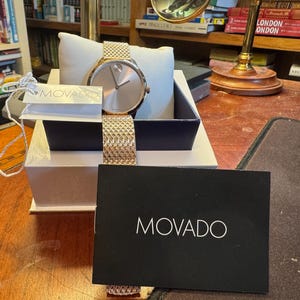 May include: A rose gold Movado watch with a mesh band, displayed in a gift box. The watch has a minimalist design with a round face and a matching rose gold band. A black card with the Movado logo is in front.