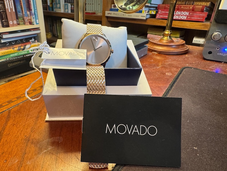 May include: A rose gold Movado watch with a mesh band, displayed in a gift box. The watch has a minimalist design with a round face and a matching rose gold band. A black card with the Movado logo is in front.