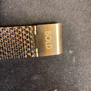 May include: Close-up of a watch band and clasp. The band is a mesh of copper-colored metal with a geometric pattern. The clasp is a matching copper color and has the word "BOLD" and "MOVADO" engraved on it.