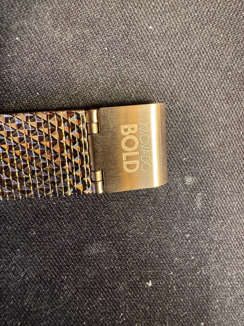 May include: Close-up of a watch band and clasp. The band is a mesh of copper-colored metal with a geometric pattern. The clasp is a matching copper color and has the word "BOLD" and "MOVADO" engraved on it.