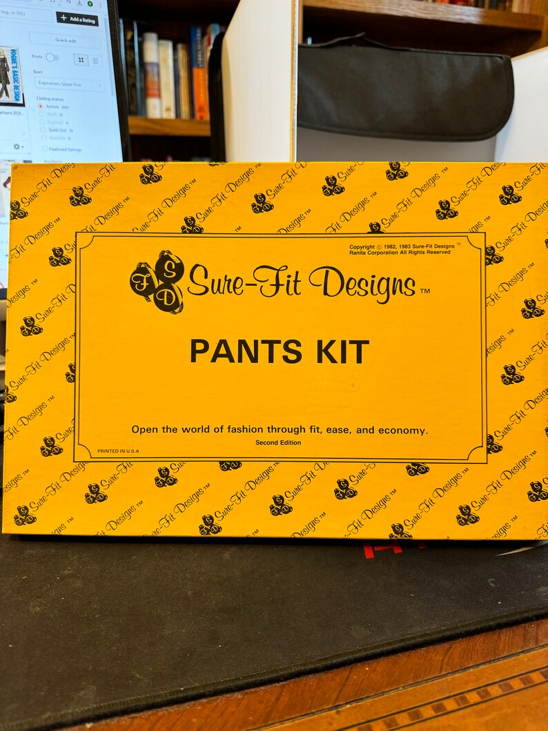K&ouml;nnte beinhalten: Eine gelbe Schachtel mit dem Text "Sure-Fit Designs" und "PANTS KIT" darauf gedruckt. Die Schachtel ist von einem Muster des Sure-Fit Designs-Logos umgeben. Der Text "Open the world of fashion through fit, ease, and economy." ist unter dem Titel gedruckt. Der Text "Second Edition" ist unter dem Text gedruckt. Der Text "Copyright 1982, 1983 Sure-Fit Designs Ranita Corporation All Rights Reserved" ist in der oberen rechten Ecke der Schachtel gedruckt.