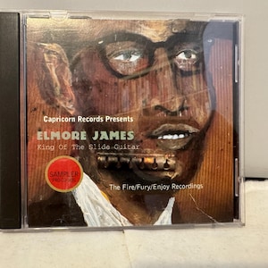 May include: CD case featuring Elmore James, the King of the Slide Guitar, with a portrait-style album cover. The artwork is a stylized, sepia-toned image of James wearing glasses. The text includes "Capricorn Records Presents" and "The Fire/Fury/Enjoy Recordings."