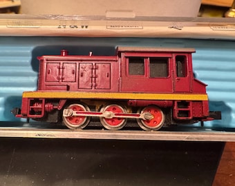 Op de afbeelding: Een kastanjebruine modeltreinlocomotief met een gele streep langs de onderkant. De motor heeft drie sets rode wielen en staat op een blauw oppervlak. De modeltrein is een speelgoed.