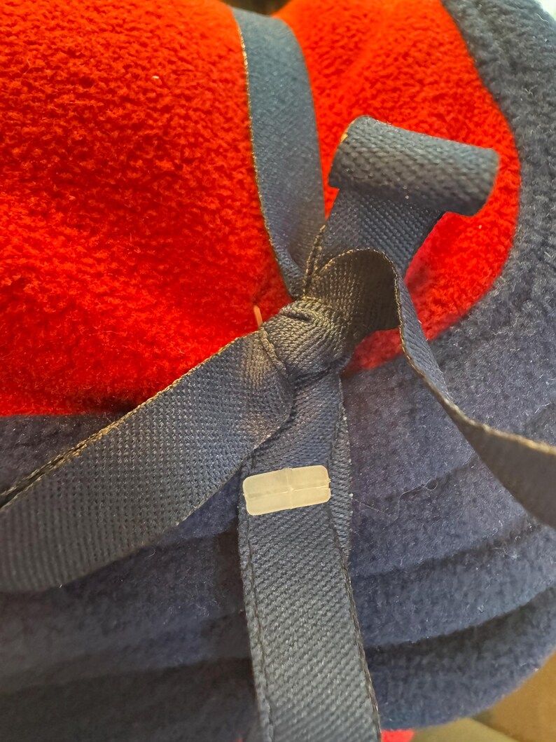 K&ouml;nnte beinhalten: Nahaufnahme einer gefalteten roten und marineblauen Fleece-Decke. Ein marineblaues Band ist mit einem Kunststoffclip um die Decke gebunden. Die Decke ist weich und flauschig, mit einer strukturierten Oberfl&auml;che.