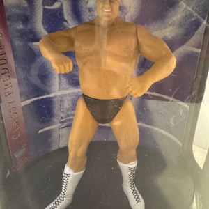 May include: A wrestling action figure of a muscular man wearing white boots and black wrestling trunks. The figure is posed with one arm raised and the other hand on his hip. The figure is in a clear plastic display case with a black base and the words "WWE" printed on the base.