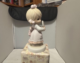 Może przedstawiać: Porcelanowa figurka młodej dziewczyny w różowej sukience, trzymającej lustro. Figurka ma blond włosy i stoi na okrągłej podstawie. Figurka znajduje się na ozdobnym pudełku z napisem "Precious Moments".