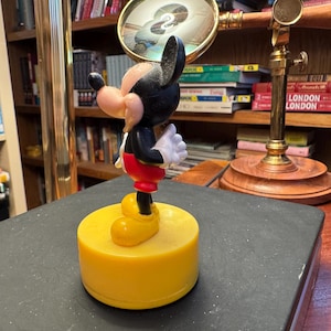 Puede incluir: Una figura de Mickey Mouse, con cuerpo negro, pantalones cortos rojos, zapatos amarillos y guantes blancos, de pie sobre una base amarilla. La figura es un personaje cl&aacute;sico de Disney, un objeto de colecci&oacute;n.