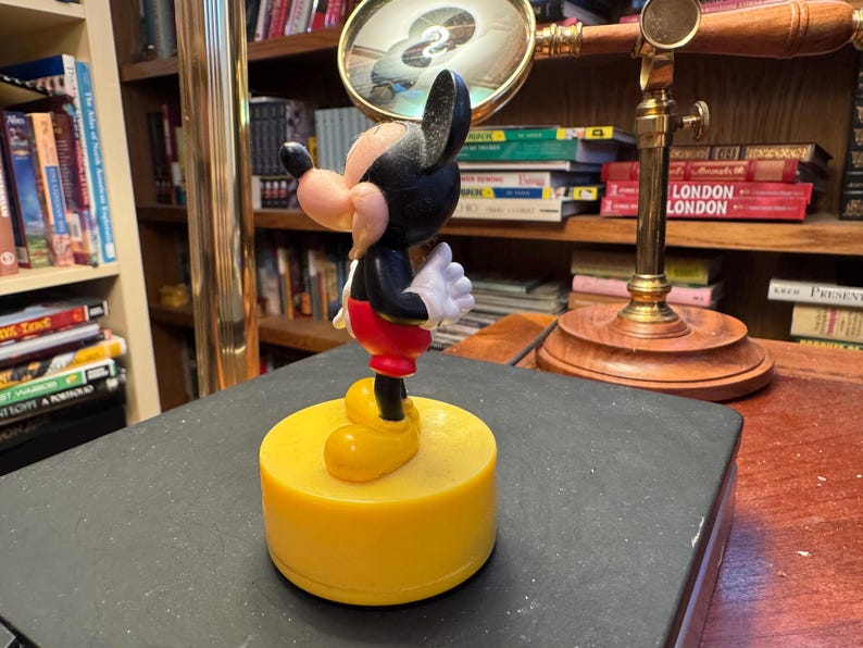 Puede incluir: Una figura de Mickey Mouse, con cuerpo negro, pantalones cortos rojos, zapatos amarillos y guantes blancos, de pie sobre una base amarilla. La figura es un personaje cl&aacute;sico de Disney, un objeto de colecci&oacute;n.