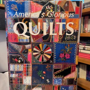 Può includere: Un libro intitolato "America's Glorious Quilts" con una copertina patchwork colorata. La copertina presenta una varietà di quadrati di tessuto nei toni del blu, rosso, rosa e nero, con dettagli floreali ricamati e il titolo in grandi lettere bianche.