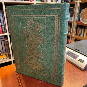 Puede incluir: Un libro de tapa dura verde con acabado texturizado y detalles florales dorados en la portada. El lomo presenta nervios y detalles en color dorado. El libro está colocado junto a una báscula digital que muestra un peso de 0,0 gramos.