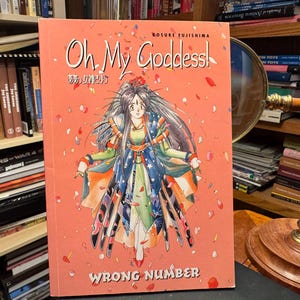 May include: A manga book titled Oh My Goddess! Wrong Number by Kosuke Fujishima with a coral orange cover featuring an illustration of a long-haired character in a layered blue and green outfit surrounded by falling pink and red flower petals.