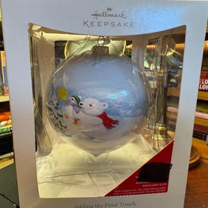 Könnte beinhalten: Ein Hallmark Keepsake Weihnachtsschmuck in einer Box. Der Schmuck ist eine große, runde, hellblaue Glaskugel mit einer Winterszene mit einem Eisbären und einem Pinguin. Der Eisbär trägt einen roten Schal. Auf der Box steht "Adding the Final Touch."