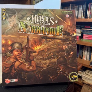 May include: The Heroes of Normandie board game box features a vibrant, illustrated war scene with soldiers in combat gear, a tank, and explosions. The title Heroes of Normandie is printed in large, stylized letters at the top, with Devil Pig Games and Iello logos.