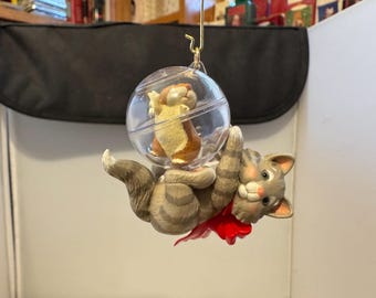 Könnte beinhalten: Ein skurriles Ornament mit einer grauen getigerten Katze mit roter Schleife, die eine durchsichtige Kugel hält. In der Kugel befindet sich ein kleines braunes und hellbraunes Eichhörnchen. Das Ornament wird von einem goldfarbenen Haken gehalten.