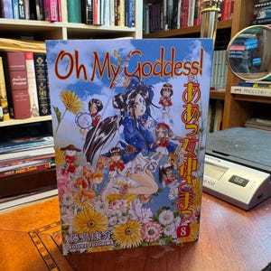 May include: A manga volume titled Oh My Goddess! volume 8 by Kosuke Fujishima. The cover features a central female character with long dark hair surrounded by smaller figures and yellow flowers against a blue sky with white clouds and Japanese text.
