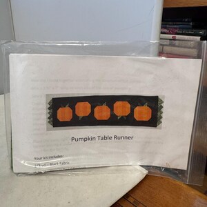 Pode incluir: Kit de artesanato para um caminho de mesa com abóboras. O design apresenta cinco abóboras laranja sobre um fundo preto. O texto "Pumpkin Table Runner" é visível na embalagem.
