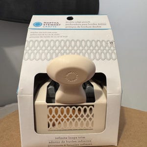 Puede incluir: Un punzón de doble borde Martha Stewart Crafts para manualidades. El paquete presenta el texto "punches one inch wide strips" y "infinite loops trim". El punzón es de color blanquecino con un mango color crema y un diseño decorativo.
