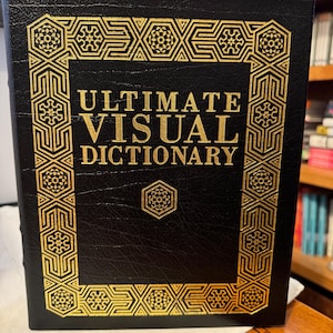 Puede incluir: Un libro de tapa dura negro con el título "ULTIMATE VISUAL DICTIONARY" en letras doradas. La portada presenta un borde decorativo dorado. El libro se muestra sobre una superficie de madera.
