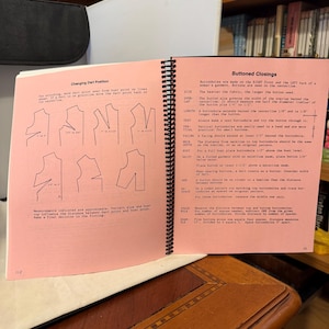 May include: Open spiral-bound sewing pattern book with pink pages. The left page shows diagrams for changing dart positions, while the right page details buttoned closings. Text and diagrams are in black. The book is open on a wooden table.