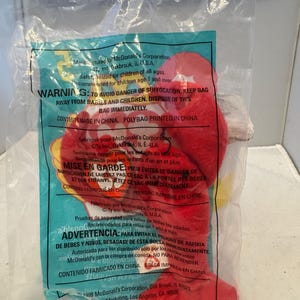 Puede incluir: Un juguete de peluche rojo dentro de una bolsa de plástico transparente. El juguete tiene un cuerpo rojo, detalles amarillos y detalles blancos. La bolsa tiene texto impreso en inglés, francés y español, incluida una advertencia de asfixia.