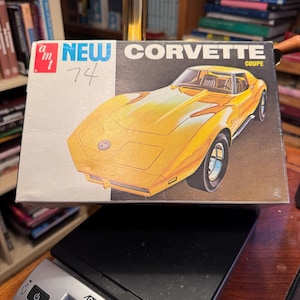 May include: A rectangular model kit box for an AMT 1974 Corvette Coupe featuring a yellow car illustration, the text "NEW" and "CORVETTE COUPE", and a handwritten "74" on the white side. The box rests on a digital scale displaying 0.15 kg.