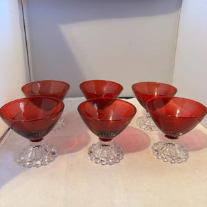 May include: Six vintage red glass champagne coupe glasses. Each glass features a ruby red bowl atop a clear, decorative stem with a beaded design. The glasses are arranged on a white surface, showcasing their elegant design.