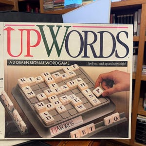 May include: UPWORDS board game box with a 3-dimensional word game. The box features the game title in red and green, with a grid of letter tiles. A hand is placing a letter tile. Ages 10 to adult.