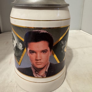 May include: A white ceramic beer stein with a portrait of Elvis Presley. The stein has a metal lid and gold trim. The image of Elvis is centered on the stein, with black and white images on either side.