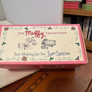May include: A pink box with the text "The Muffy Collection" and "Jam Making Jar Set Jam Session." The box has illustrations of jam jars and labels. The box is on a wooden surface.