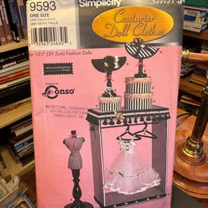 May include: A Simplicity Series 4 Couturier Doll Clothes pattern for 15 ½ inch fashion dolls. The pink package features illustrations of doll clothes, a dress form, and a clothing rack. The text includes "One Size" and "Heigl Nordstrom Designs."