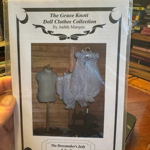May include: A package featuring a doll clothes collection by Judith Marquis. The image inside shows a miniature dress, bloomers, and a dress form. The text reads "The Grace Knott Doll Clothes Collection" and "The Dressmaker's Judy & Undies."