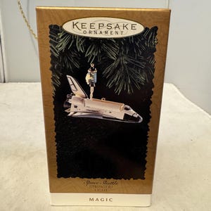 May include: A gold-colored box with a Christmas ornament inside. The ornament is a silver space shuttle with a small astronaut figure on top. The box has the words "Keepsake Ornament" and "Space Shuttle Stringer! Light Magic" printed on it.