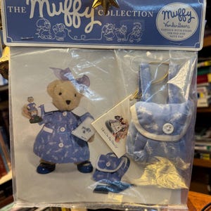 Può includere: Un set della collezione Muffy VanderBeare con una bambola orsetto in un vestito blu, scarpe e fiocco. Il set include uno zaino abbinato e un blocco note con timbro e tampone per inchiostro. La confezione recita "The Muffy Collection."