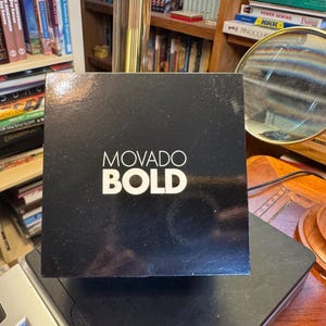 May include: A black box with the words "MOVADO BOLD" in white lettering. The box is sitting on a black surface, with a bookshelf and a magnifying glass in the background.