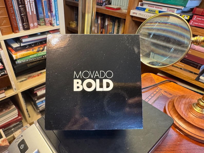 May include: A black box with the words "MOVADO BOLD" in white lettering. The box is sitting on a black surface, with a bookshelf and a magnifying glass in the background.