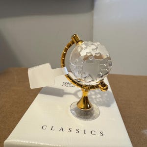 Peut inclure: Une figurine de globe terrestre en cristal avec un support et un cadre dorés. Le globe est transparent et facetté, reposant sur une base dorée. La figurine est posée sur une boîte blanche avec le mot "CLASSICS" imprimé.