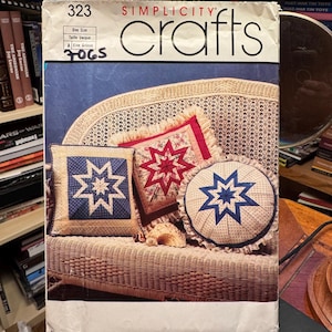 Puede incluir: Un sobre de patrón Simplicity Crafts número 323 para cojines de estrella, un agarrador de ollas y un tapiz. La portada muestra tres cojines decorativos con diseños de estrellas en colores azul, rojo y crema sobre una superficie de mimbre.