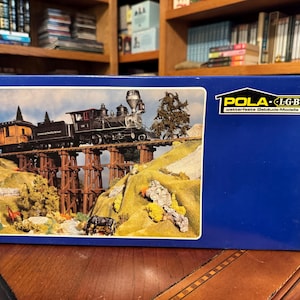 Puede incluir: Una caja azul que muestra una escena de tren en miniatura. El tren, negro y marrón, cruza un puente de madera sobre un paisaje con colinas, árboles y un arroyo. Se puede ver el logotipo "POLA-LGB".