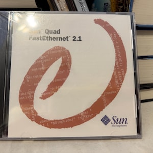May include: A CD case for Sun Quad FastEthernet 2.1 software. The cover is white with a large orange "e" shape and text. The Sun Microsystems logo is in the lower right corner. The CD case is in a clear plastic case.