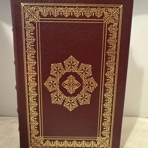 Pode incluir: Um livro encadernado em couro bordô com uma borda decorativa dourada e um desenho dourado no centro da capa.