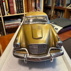 May include: A gold-coloured scale model car featuring a prominent chrome front bumper, a detailed mesh grille, and dual round headlights. The vehicle has a bonnet with ventilation slats, a windscreen with wipers, and side-mounted mirrors on the wings.