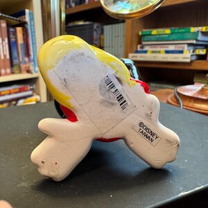 Pu&ograve; includere: Statuetta in ceramica di un personaggio dei cartoni animati, con capelli gialli, abito rosso e guanti bianchi. La statuetta &egrave; contrassegnata "&copy;DISNEY TAIWAN" sulla base. Su una superficie nera.